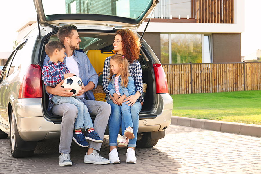 Personal Insurance - Young Family with Children Sitting in Their Car Trunk Near Their Family Home on a Summer Day